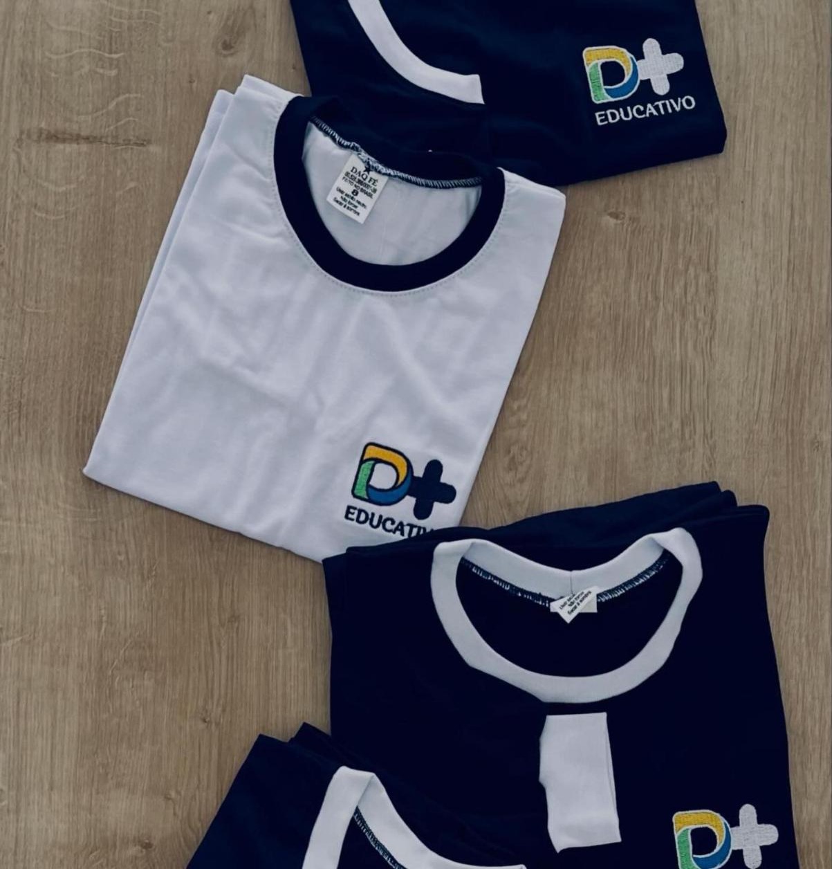 Camisetas branca e azul marinho do D+ Educativo dobradas sobre madeira, com o bordado da escola