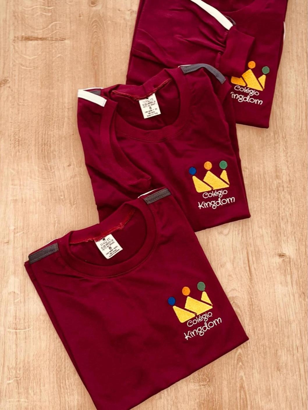 Três camisetas bordô do Colégio Kingdom dobradas sobre madeira, com o bordado da coroa e a etiqueta Daq Fé