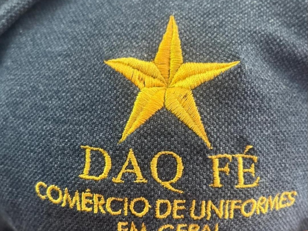 Bordado dourado da marca Daq Fé em piquê cinza azulado: estrela de cinco pontas e o nome Comércio de Uniformes em Geral