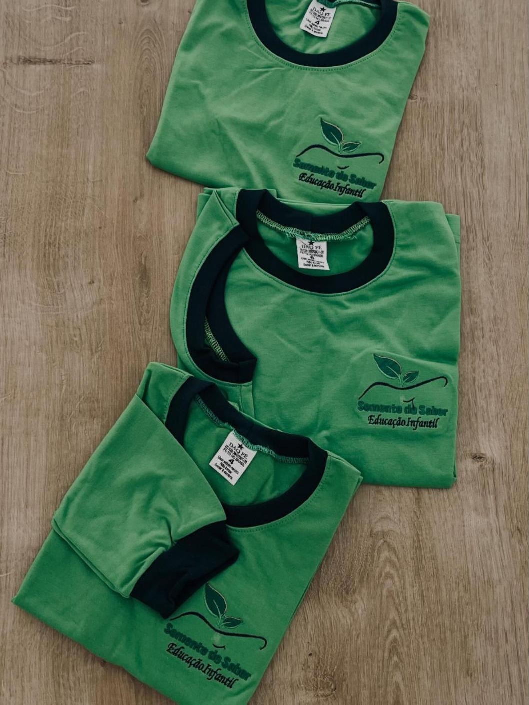 Três camisetas verdes da Semente do Saber Educação Infantil, com gola azul marinho e etiqueta Daq Fé visível